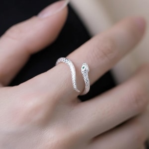 May include: A silver snake ring with a textured pattern. The snake's head is adorned with tiny, green gemstones for eyes. The ring coils around the finger, offering a distinctive and fashionable accessory.