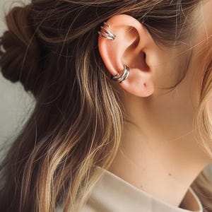 Puede incluir: Juego de joyas de puño de oreja plateado. La imagen muestra un primer plano de una oreja adornada con múltiples puños de oreja plateados. Los puños están diseñados con una estética elegante y minimalista, con un acabado liso y pulido. Los puños de oreja están diseñados para envolver el cartílago de la oreja.