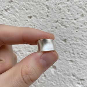 May include: A silver ring with a wide, curved band. The ring has a matte finish and is held between two fingers against a textured white background. The ring's design is simple and modern, suitable for everyday wear.