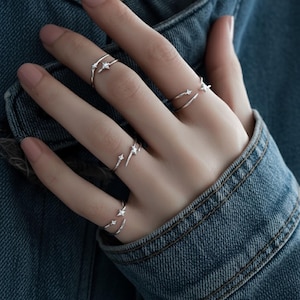 Könnte beinhalten: Nahaufnahme einer Hand mit mehreren silbernen Ringen. Jeder Ring hat ein zartes Sterndesign. Die Ringe werden an mehreren Fingern getragen und erzeugen einen geschichteten Effekt. Der Hintergrund ist eine Jeansjacke.