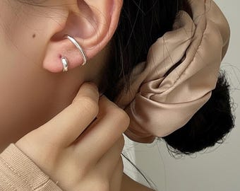 Puede incluir: Puño de oreja plateado y pendiente pequeño. El puño de oreja se curva alrededor de la parte superior de la oreja, mientras que el pendiente es un diseño pequeño y redondeado. La joya se lleva en la oreja de una persona, con una goma para el pelo beige en el fondo.