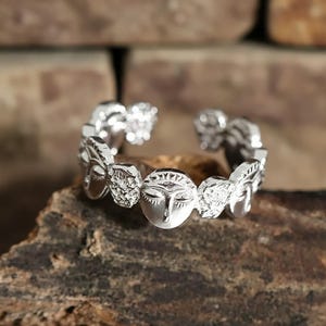 May include: A silver-colored ring with a detailed design featuring stylized faces and decorative elements. The ring is set against a natural, textured background, highlighting its intricate craftsmanship and unique aesthetic.