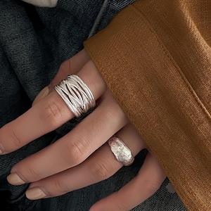 May include: Two silver rings are shown on a hand. One is a wide band made of interwoven strands. The other ring has a textured, organic design. The background includes denim and a brown garment.