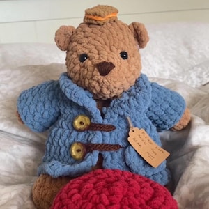Könnte beinhalten: Ein gehäkelter brauner Bär mit einem blauen Mantel mit Holzknöpfen und einem kleinen Hut. Vor dem Bären liegt ein roter Hut. Der Text "BEAR CROCHET PATTERN" steht oben im Bild.