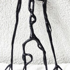Peut inclure: Une sculpture tridimensionnelle noire et brillante dotée d'une tête arrondie, de bras allongés pointant vers le bas et d'un torse élancé aux formes interconnectées. La figurine présente une surface lisse et réfléchissante sur des pieds fins.