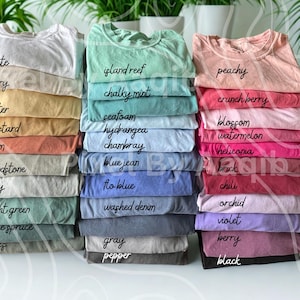 May include: Stacks of folded t-shirts in various colors, including white, pink, and blue. Each shirt is labeled with a color name in a handwritten font. The shirts are neatly arranged, showcasing a range of hues.