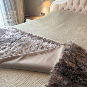 May include: A bed with a beige textured bedspread and a decorative faux fur throw in shades of brown and tan. The headboard is white with a tufted, beige upholstered design. The room has a tropical wallpaper.