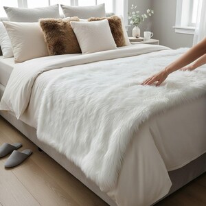 May include: A bed is made with white bedding, including pillows and a fluffy white faux fur throw blanket. The bed is in a well-lit room with a neutral colour palette. Grey slippers are on the floor.
