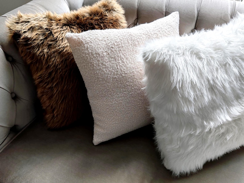 Pu&ograve; includere: Tre cuscini decorativi su un divano grigio. Un cuscino &egrave; in finta pelliccia marrone, uno &egrave; bianco con una superficie strutturata e l'altro &egrave; in finta pelliccia bianca. Il divano ha un design trapuntato.