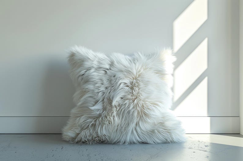 Pu&ograve; includere: Un cuscino quadrato e soffice bianco con lunghi peli strutturati. Il cuscino &egrave; posizionato contro un muro bianco e poggia su un pavimento grigio chiaro. La luce del sole proietta ombre sul muro.
