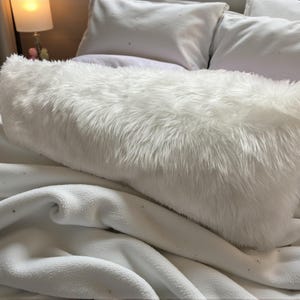 Pu&ograve; includere: Un cuscino rettangolare bianco e soffice con peli lunghi e morbidi. Il cuscino &egrave; posto su una coperta bianca ed &egrave; circondato da altri cuscini bianchi, creando un'atmosfera accogliente e invitante. La biancheria da letto &egrave; di colore neutro.