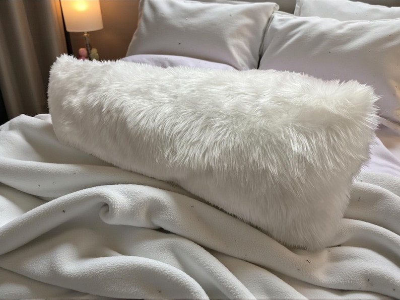 Pu&ograve; includere: Un cuscino rettangolare bianco e soffice con peli lunghi e morbidi. Il cuscino &egrave; posto su una coperta bianca ed &egrave; circondato da altri cuscini bianchi, creando un'atmosfera accogliente e invitante. La biancheria da letto &egrave; di colore neutro.