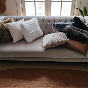 Pu&ograve; includere: Un divano grigio trapuntato &egrave; adornato da una variet&agrave; di cuscini decorativi. I cuscini presentano finta pelliccia nei toni del marrone, bianco, grigio e nero. &Egrave; presente anche un lungo cuscino rettangolare in pelle marrone.