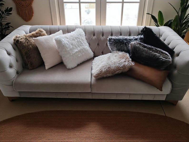 Pu&ograve; includere: Un divano grigio trapuntato &egrave; adornato da una variet&agrave; di cuscini decorativi. I cuscini presentano finta pelliccia nei toni del marrone, bianco, grigio e nero. &Egrave; presente anche un lungo cuscino rettangolare in pelle marrone.