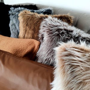 Pu&ograve; includere: Una collezione di cuscini decorativi in varie trame e colori. I cuscini includono finta pelliccia in nero, grigio, marrone e beige, un cuscino in sherpa marrone chiaro e un cuscino in pelle marrone. Sono disposti su un divano grigio trapuntato.