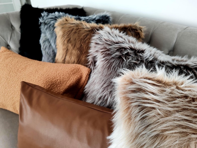 Pu&ograve; includere: Una collezione di cuscini decorativi in varie trame e colori. I cuscini includono finta pelliccia in nero, grigio, marrone e beige, un cuscino in sherpa marrone chiaro e un cuscino in pelle marrone. Sono disposti su un divano grigio trapuntato.