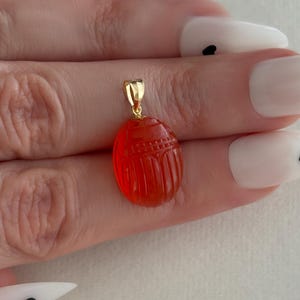 May include: A vibrant red, oval-shaped scarab beetle pendant carved from translucent stone. The pendant features a detailed, segmented insect design on its surface and is topped with a polished gold-toned metal bail for attachment to a chain.