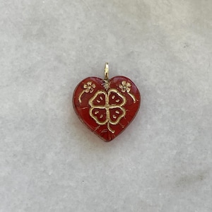 May include: A heart-shaped pendant with a red enamel finish and gold-colored accents. The pendant features a four-leaf clover design in the center and floral details on the top corners. The bail is gold-colored.