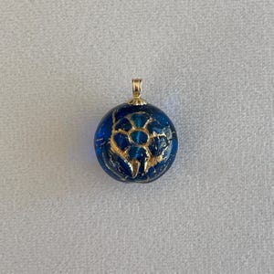 May include: A round, blue pendant with a gold-colored bail. The pendant features a floral design in gold and a textured, crackled appearance. The pendant is a jewelry item.