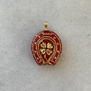 May include: A gold-colored pendant with a red horseshoe design and a four-leaf clover in the center. The pendant has a textured surface and a small loop at the top for a chain. The clover is gold-colored.