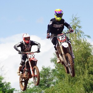 Könnte beinhalten: Zwei Motocross-Fahrer in der Luft vor blauem Himmel. Einer trägt Schwarz-Weiß mit der Nummer 117 auf dem Motorrad. Der andere trägt Schwarz mit "SHIFT" und der Nummer 13 auf dem Motorrad. Die Motorräder sind rot und braun.