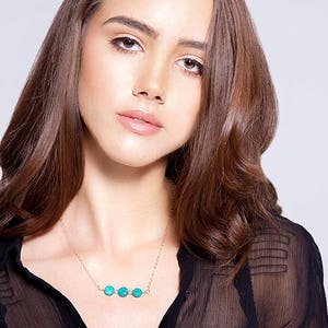 Puede incluir: Una mujer con una blusa negra transparente con un collar de cadena dorada con cinco cuentas redondas de color turquesa.