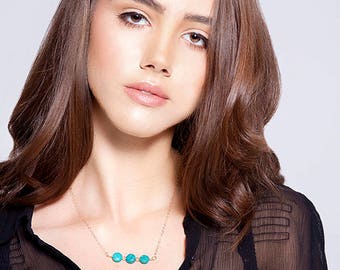 May include: A woman wearing a black sheer top with a gold chain necklace featuring five turquoise round beads.