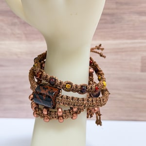 May include: Three macrame bracelets in shades of brown and copper are stacked on a white surface. The bracelets feature a variety of beads in red, yellow, and brown, with a rectangular dark stone accent.