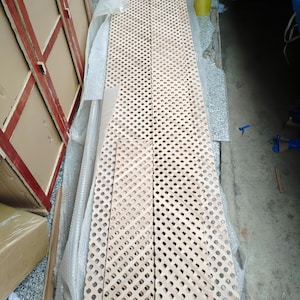 a long, perforated wooden plank lying on the ground, surrounded by cardboard boxes and other materials, suggesting it is being used for storage or construction purposes.