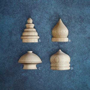 May include: Four wooden finials in various shapes, including a tiered pagoda-like design, a rounded dome, and a mushroom-shaped form. The finials are made of light-colored wood and are displayed against a textured blue background.