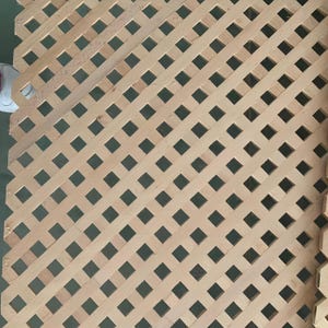 May include: Wooden lattice panel with a diamond pattern. The light brown wood is arranged in a diagonal grid, creating a series of square openings. The panel is likely for decorative or structural use.