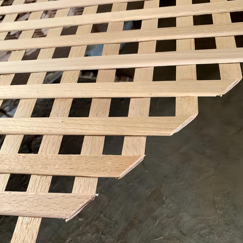 May include: Close-up of a wooden lattice structure. The lattice is made of light-colored wood, with a grid pattern of rectangular openings. The wood grain is visible, and the edges are cleanly cut. The background is a muted gray.