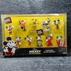 May include: A collectible deluxe figure set featuring Mickey Mouse in various poses. The set includes a golden Mickey figure and other figures in red, black, yellow, and white. The packaging has the text "90 Years of Magic".