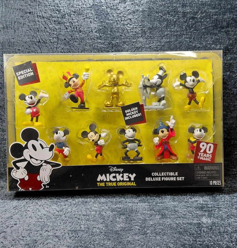 May include: A collectible deluxe figure set featuring Mickey Mouse in various poses. The set includes a golden Mickey figure and other figures in red, black, yellow, and white. The packaging has the text "90 Years of Magic".