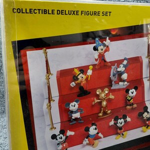 May include: A collectible deluxe figure set featuring various Mickey Mouse figurines in different poses and outfits. The figures are displayed on a red carpet-like surface, with the text "COLLECTIBLE DELUXE FIGURE SET" visible on the packaging.