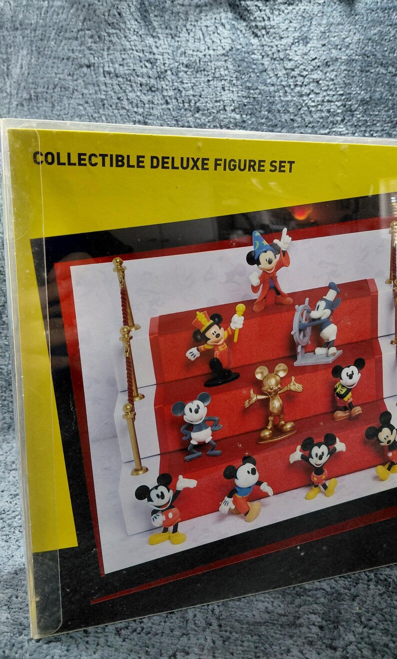 May include: A collectible deluxe figure set featuring various Mickey Mouse figurines in different poses and outfits. The figures are displayed on a red carpet-like surface, with the text "COLLECTIBLE DELUXE FIGURE SET" visible on the packaging.