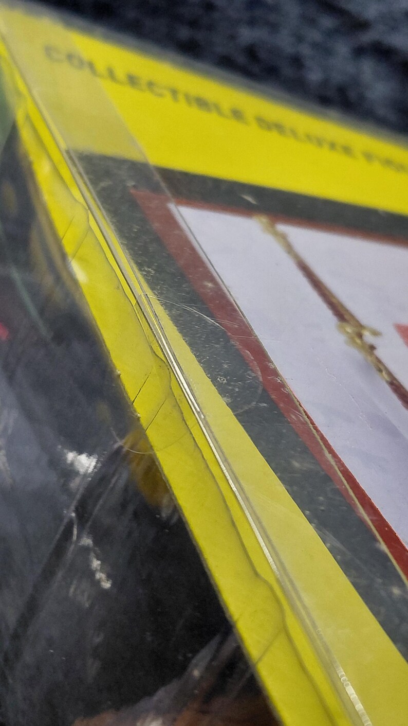 May include: A packaged collectible item with a yellow border and the words "COLLECTIBLE DELUXE FIGURE" visible. The packaging is clear plastic, revealing a glimpse of the figure inside. The overall color scheme includes yellow, red, and black.