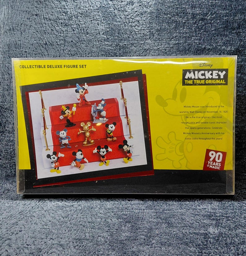 May include: Collectible Deluxe Figure Set featuring Mickey Mouse characters. The set includes various Mickey Mouse figurines in different poses, displayed on a red platform. The packaging is yellow with the text "MICKEY THE TRUE ORIGINAL" and "90 YEARS MAGIC".