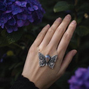 Puede incluir: Un collar con colgante de mariposa plateada con una piedra iridiscente en forma de l&aacute;grima en el centro. Las alas de la mariposa est&aacute;n detalladas con patrones florales y de filigrana. El collar se muestra sobre un fondo de flores y follaje morado.