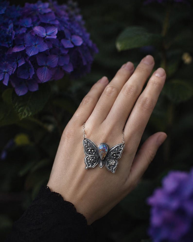 Puede incluir: Un collar con colgante de mariposa plateada con una piedra iridiscente en forma de l&aacute;grima en el centro. Las alas de la mariposa est&aacute;n detalladas con patrones florales y de filigrana. El collar se muestra sobre un fondo de flores y follaje morado.