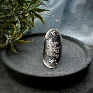 May include: A silver-toned ring with a bear design, a crescent moon, and a star. The bear is reaching towards the moon. The ring is set against a dark oval background with a textured appearance. The ring is on a dark gray, round tray.