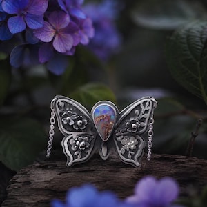 Puede incluir: Un colgante de mariposa plateada con intrincados detalles florales y una piedra de &oacute;palo central. El colgante tiene delicados detalles de cadena y se encuentra sobre un fondo borroso de flores y follaje morado.