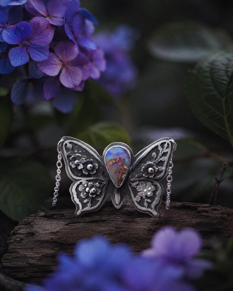 Puede incluir: Un colgante de mariposa plateada con intrincados detalles florales y una piedra de &oacute;palo central. El colgante tiene delicados detalles de cadena y se encuentra sobre un fondo borroso de flores y follaje morado.