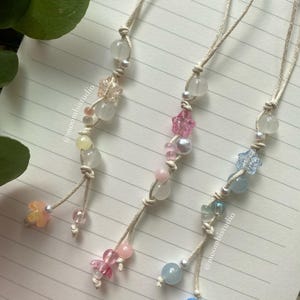 May include: Three beaded charms with a variety of pastel-colored beads and flower-shaped accents. Each charm is strung on a beige cord and features a loop at the top. The charms are displayed on a lined notebook.