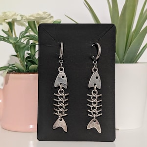 May include: A pair of silver-coloured fish skeleton earrings. Each earring has a fish bone design with a chain connecting the pieces. The earrings are displayed on a black card, with plants in the background.