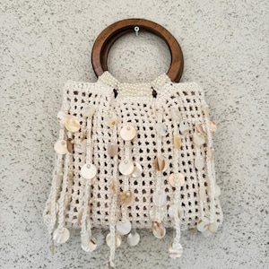 May include: A cream-colored crocheted handbag with a wooden handle. The bag is adorned with dangling shell accents and small beads. The handle is a dark brown, and the bag is hanging against a textured gray wall.