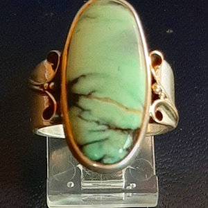 Puede incluir: Un anillo de plata con una piedra grande, ovalada, de color verde claro con vetas oscuras. La piedra está engastada en un bisel dorado. El anillo tiene detalles decorativos de plata en los laterales.
