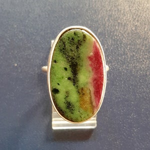 Op de afbeelding: Ovale ring met een grote edelsteen in groen, rood en geel. De ring heeft een zilveren band. De sierlijke ring staat tegen een blauwe achtergrond.