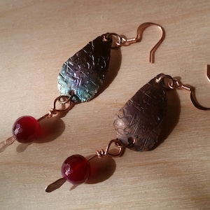 May include: A pair of earrings with copper wire and red beads. The earrings feature a teardrop-shaped copper pendant with a textured surface.