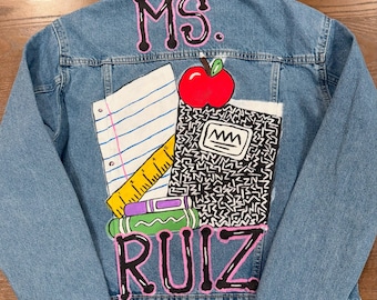 May include: Light blue denim jacket with a colorful school-themed design. The back features the text "MS. RUIZ" in pink lettering, along with illustrations of an apple, notebook, ruler, and pencils. A unique, personalized teacher gift.